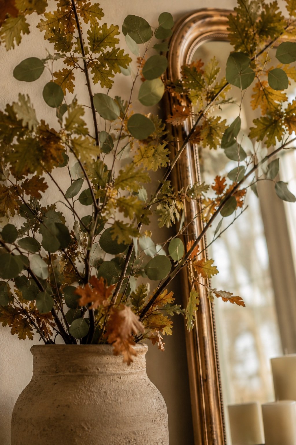 Tall cut oak and eucalyptus branches in a stoneware vase reaching above the mantel mirror