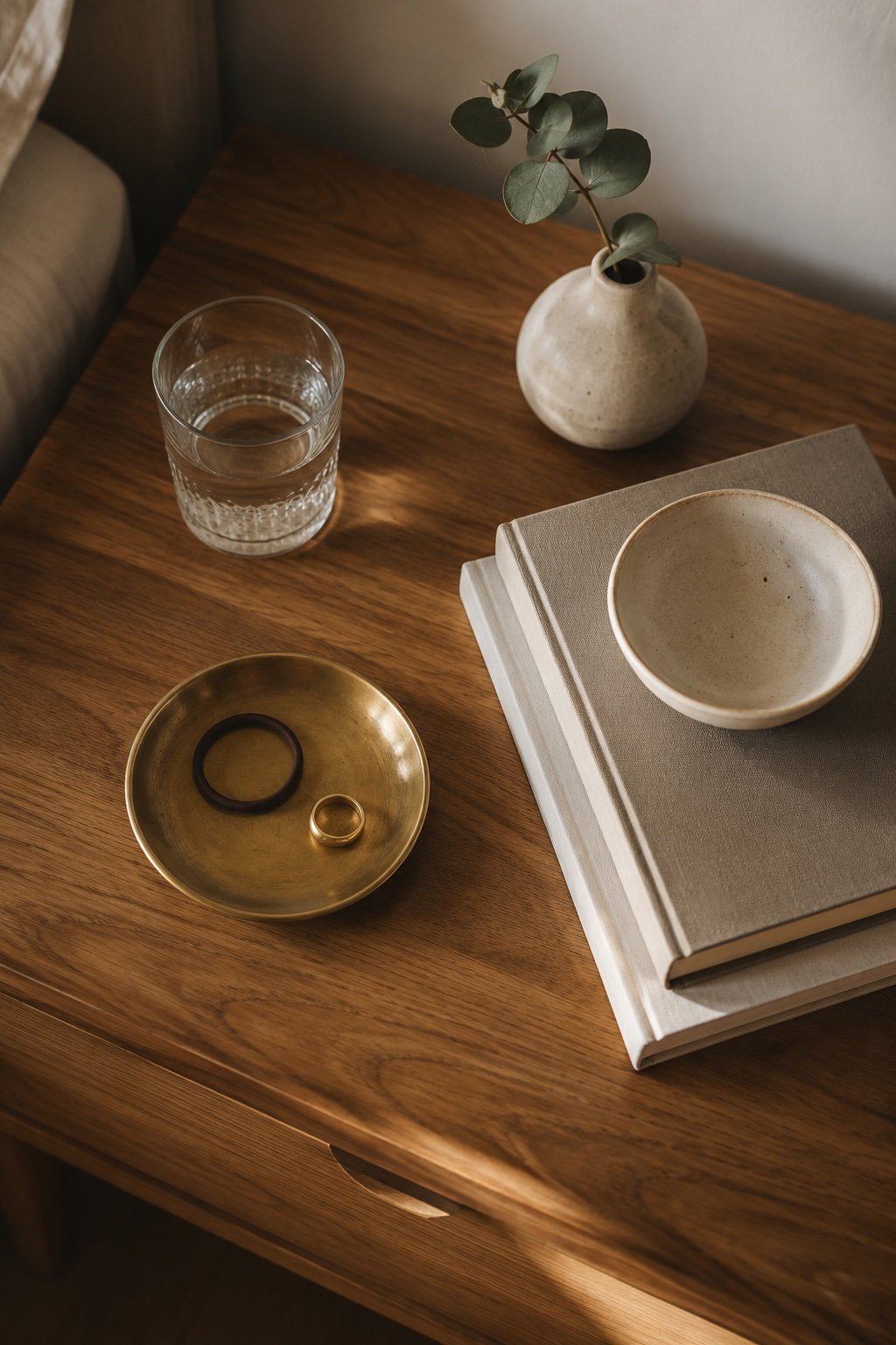 Nightstand surface detail with a brass dish, a flat book stack topped by a small bowl and a bud vase